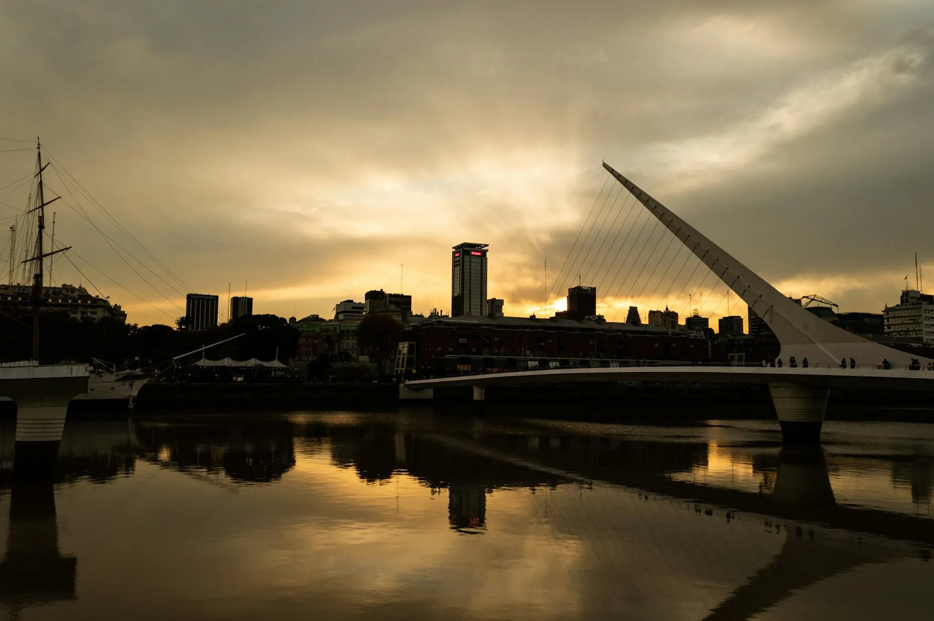 Silhouette of a city skyline with a modern bridge at sunset reflecting in calm waters.