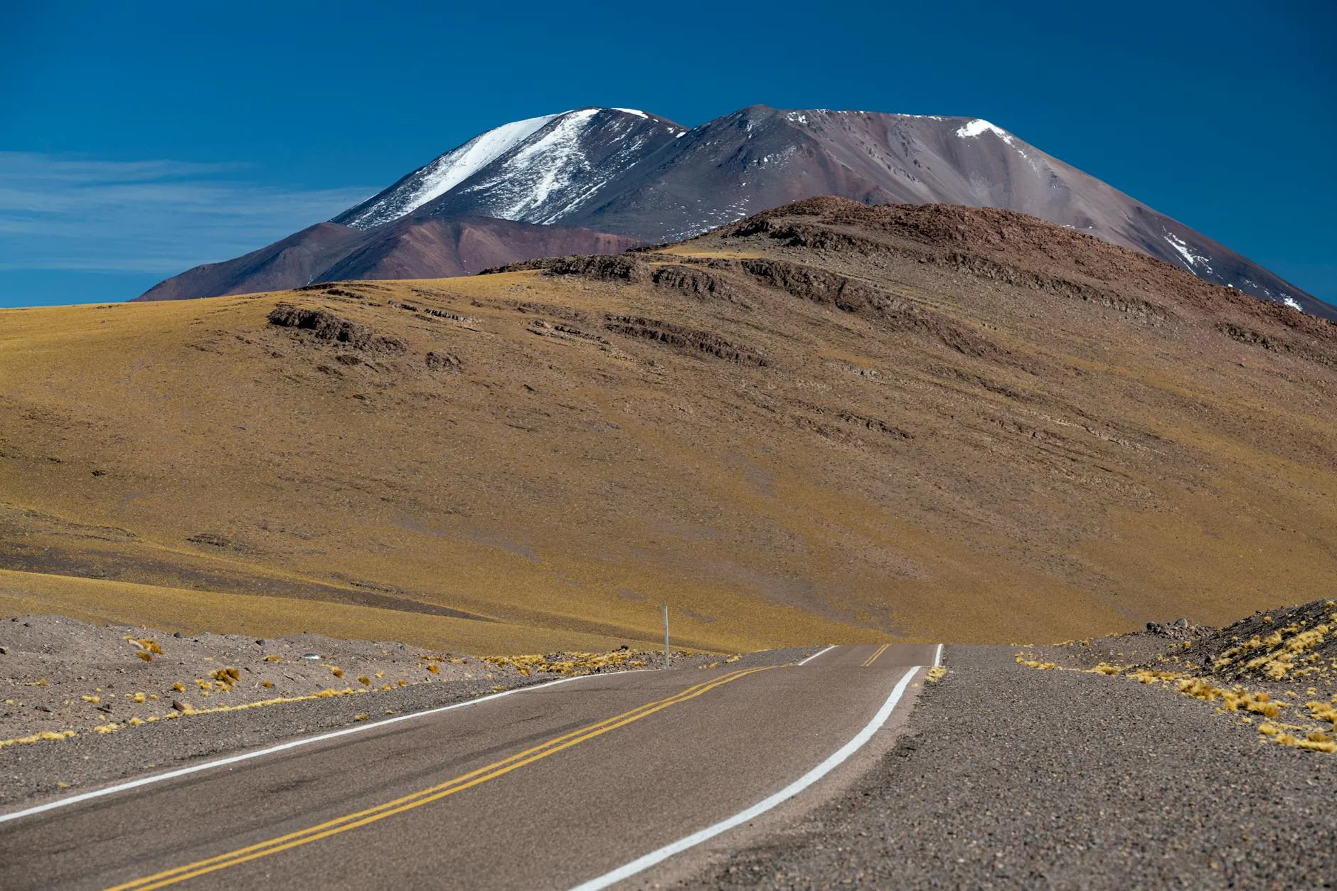 Desolate road leading to snow-capped mountains under clear blue skies.