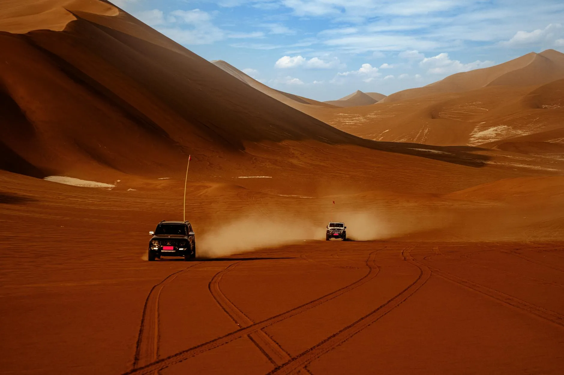 Two SUVs driving through an expansive desert with sand dunes under a sunny sky.