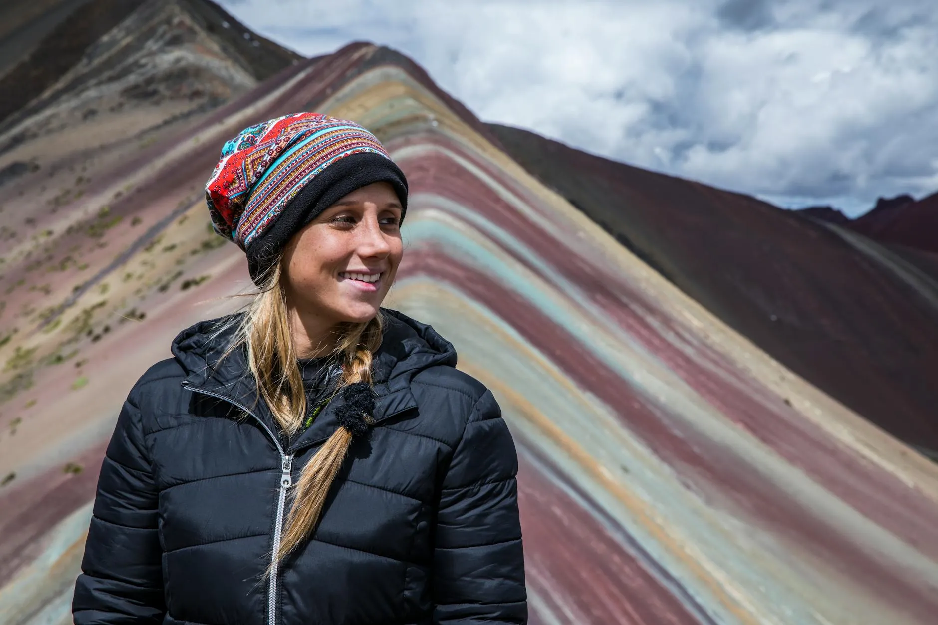 Smiling woman in a warm jacket poses at the vibrant Vinicunca Rainbow Mountain in Peru, showcasing stunning colors.